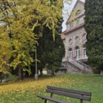 V zábere je lavička obklopená nádhernou zeleňou. V pozadí hlavná budova z boku.